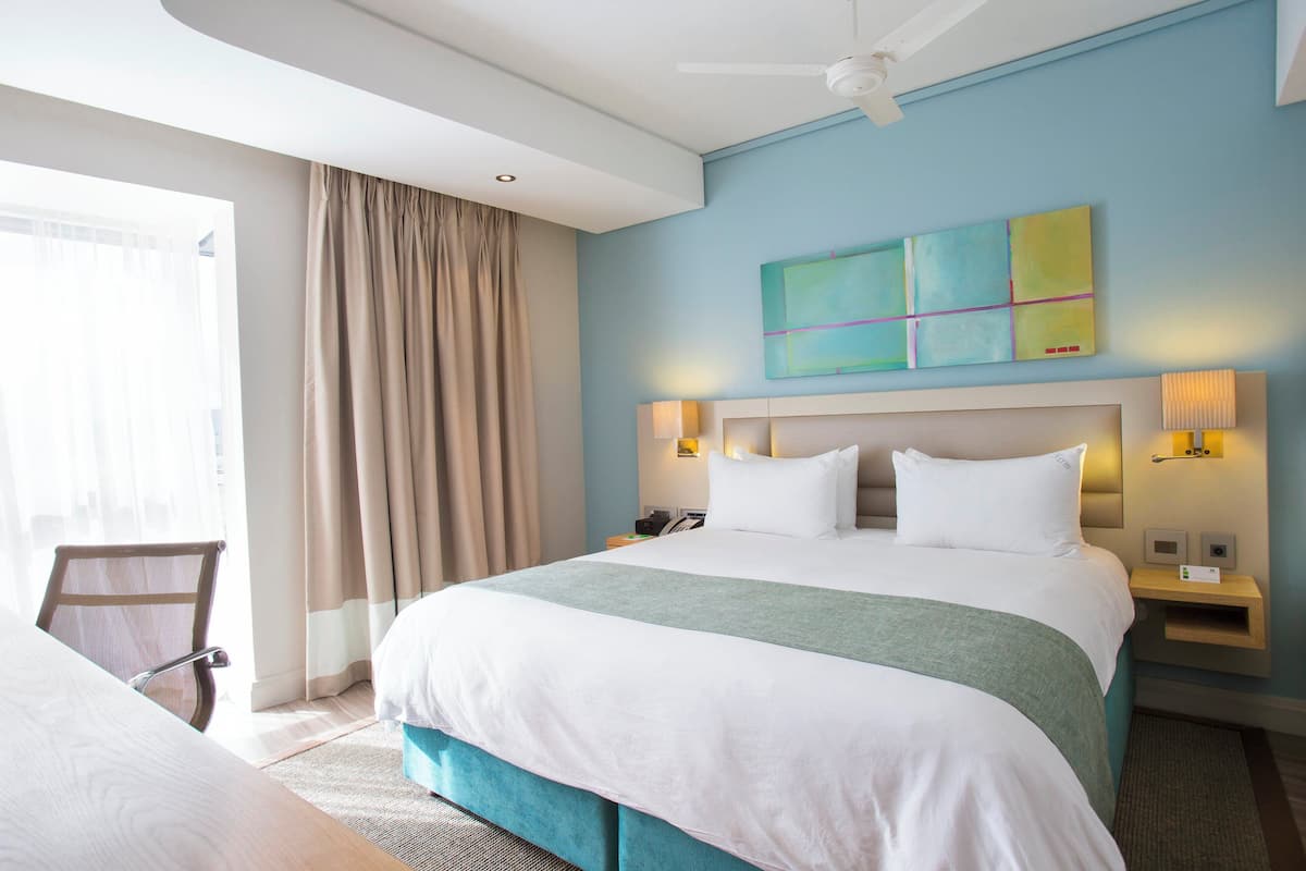 Une chambre d’hôtel avec un grand lit, un bureau et une chaise. Il y a une fenêtre avec des rideaux, un ventilateur de plafond et un mur orné d’un tableau.