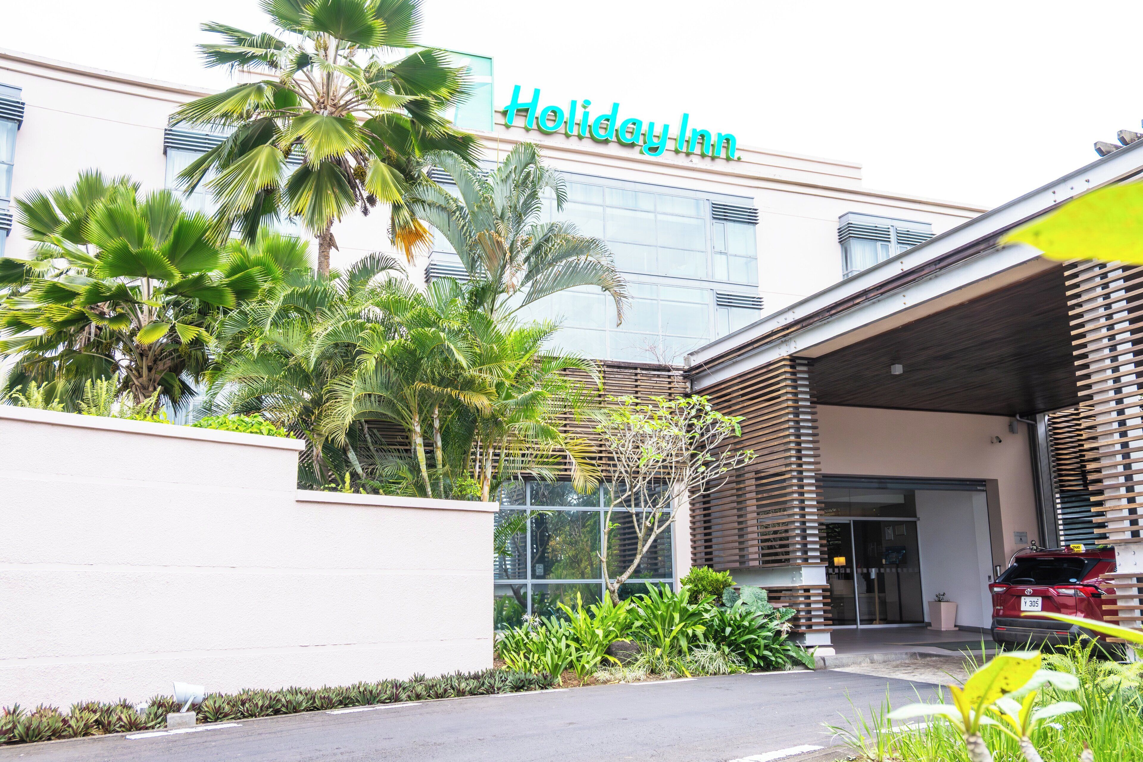 Un hotel Holiday Inn con fachada moderna, palmeras y un coche rojo estacionado al frente.