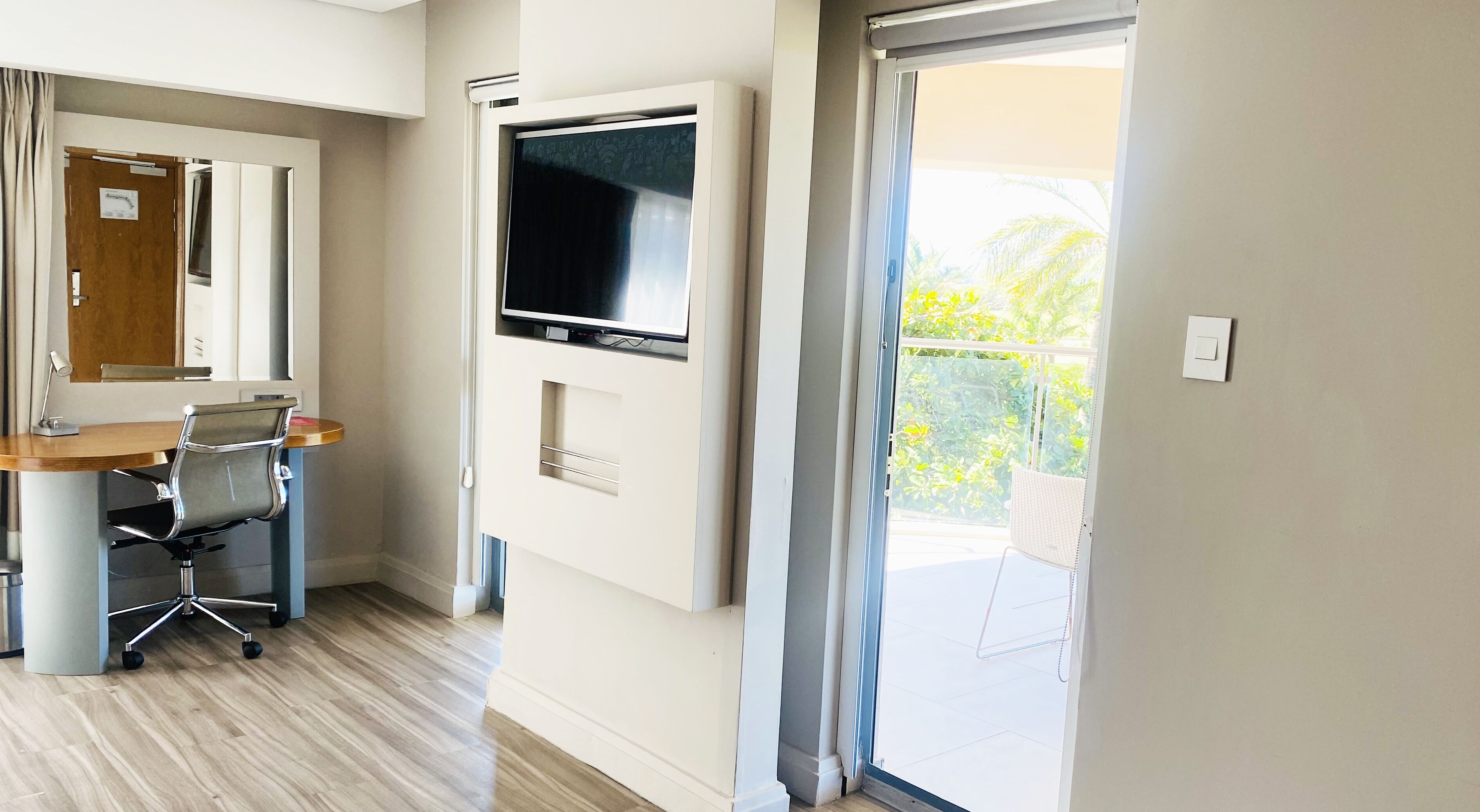 Habitació d'hotel moderna amb escriptori de fusta, una cadira, televisió plana penjada a la paret i porta corredissa de vidre que dona a un balcó amb vistes a vegetació.