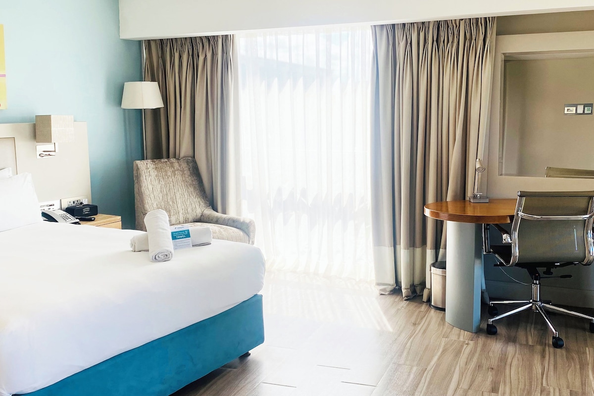 A hotel room with a large bed, a desk with a chair, a lamp, and a view of the outdoors through the window.