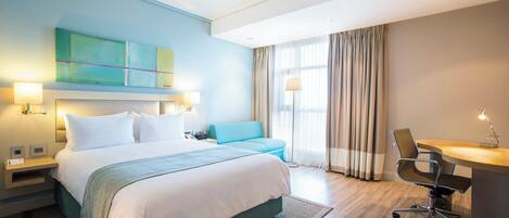 A hotel room with a bed, a desk, a chair, a sofa, a ceiling fan, and a window with curtains.