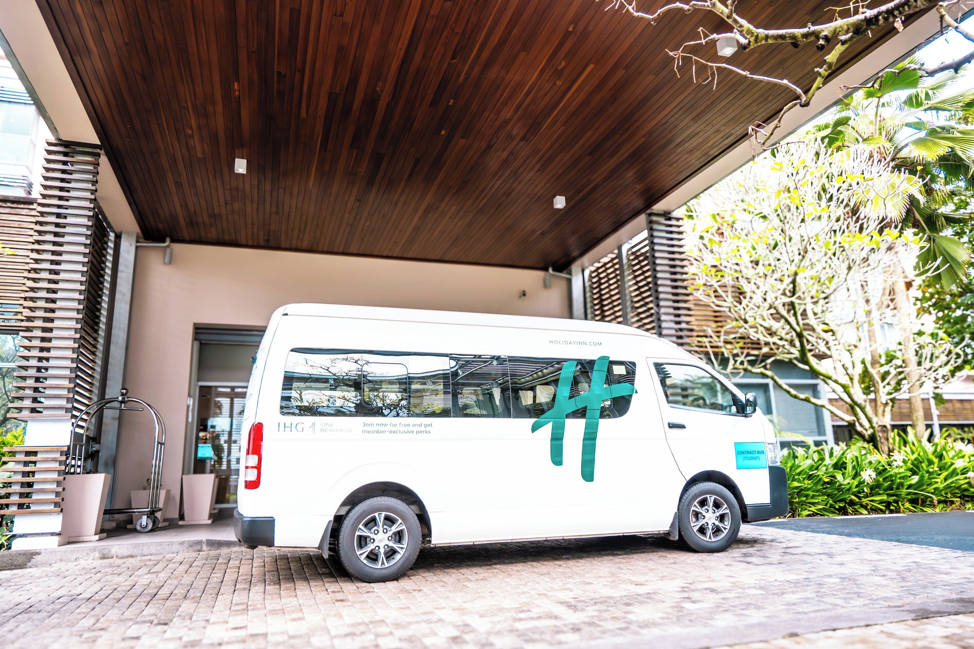 Un fourgon blanc était garé sous une avancée en bois, avec un panneau indiquant « Autobus de transport scolaire », à proximité d’un bâtiment doté d’une grille métallique et agrémenté de verdure.