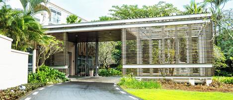 A modern building with a covered entrance, lush greenery, and a paved driveway.