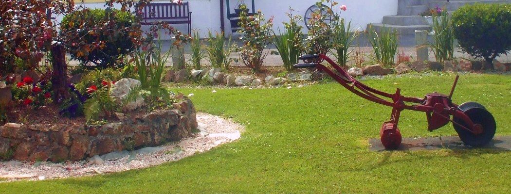 Un jardín con una herramienta roja de jardinería, un muro de piedra y diversas plantas.