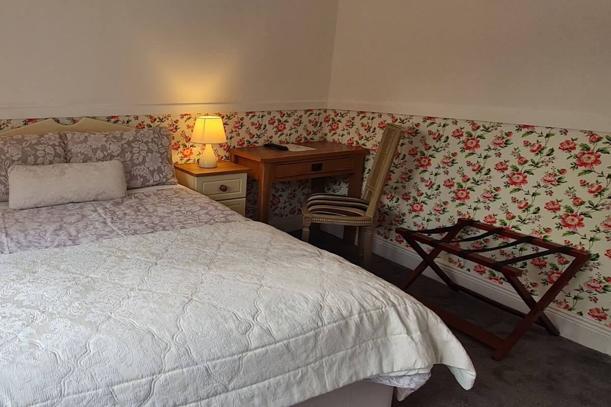 A bedroom with a bed, two bedside tables with lamps, a wooden desk, a chair, and floral wallpaper.