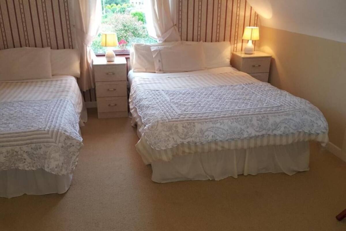 A bedroom with two beds, each with a white bedspread and a matching headboard. There are bedside tables with lamps and a window with curtains.