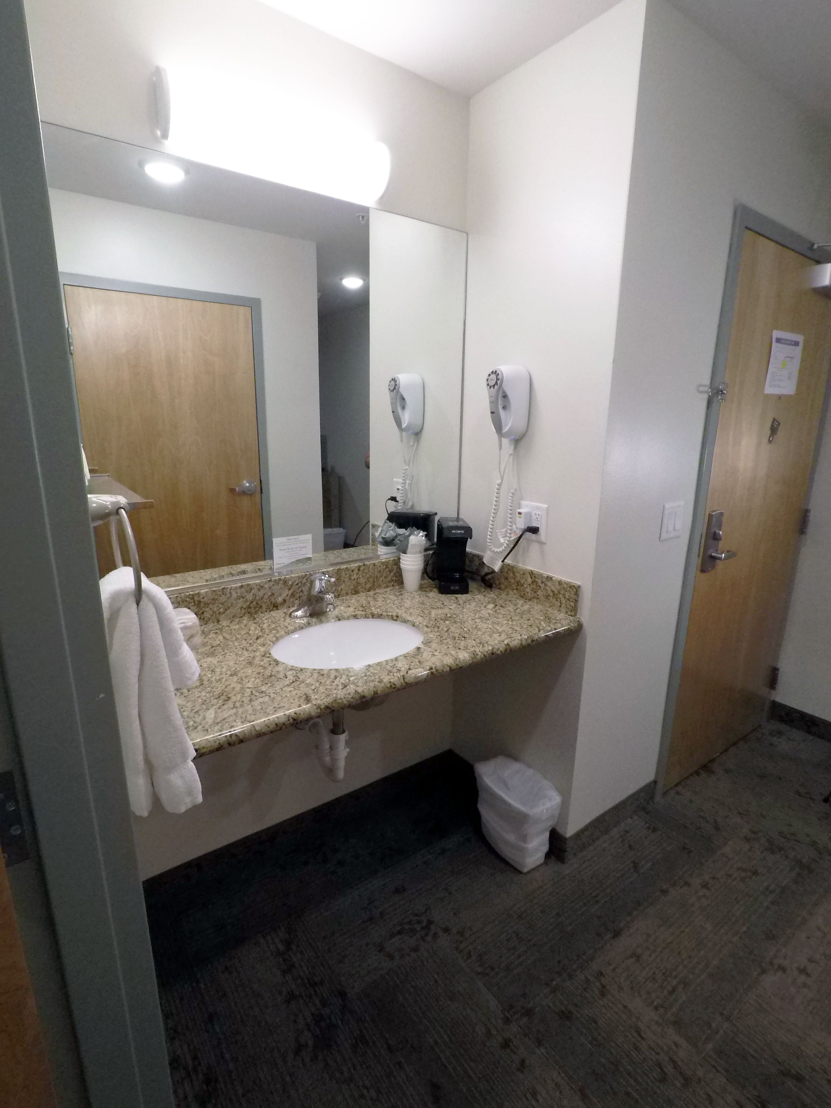 A hotel bathroom with a granite countertop sink, a mirror, two hair dryers, a trash bin, and a wooden door.