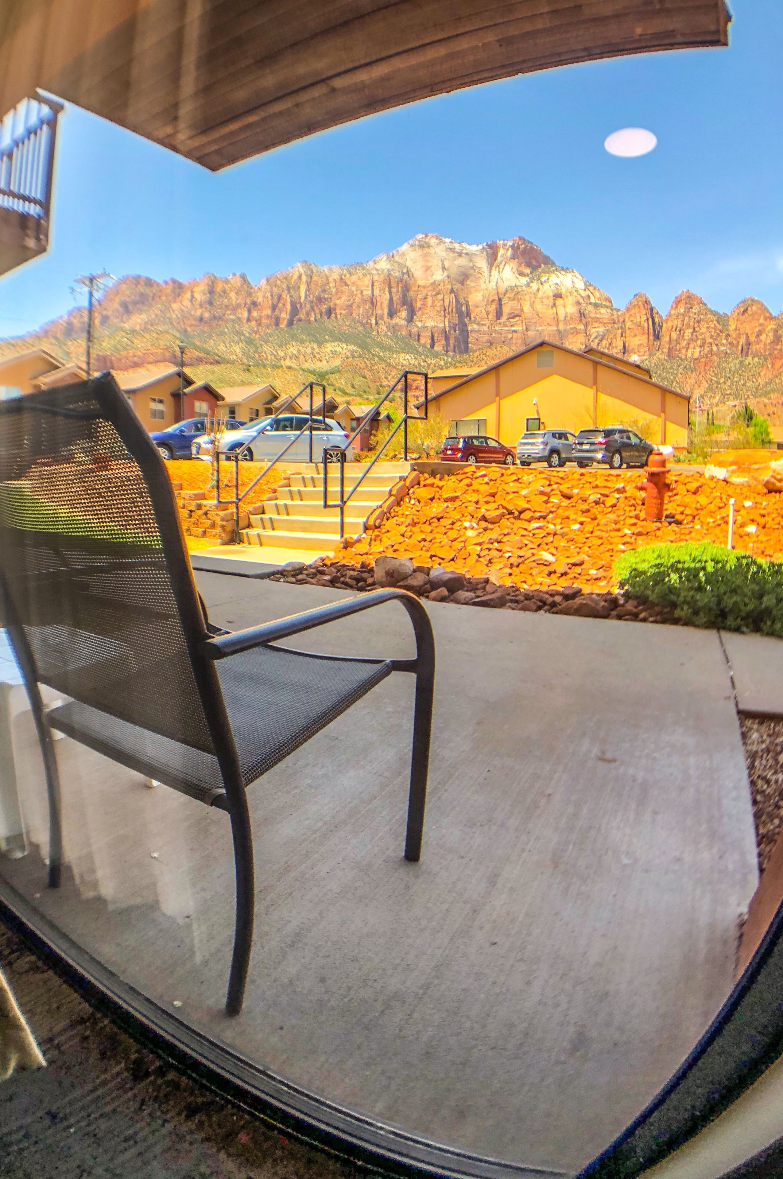 A view from a window with a metal chair, a building with a yellow facade, and a mountain range in the background.