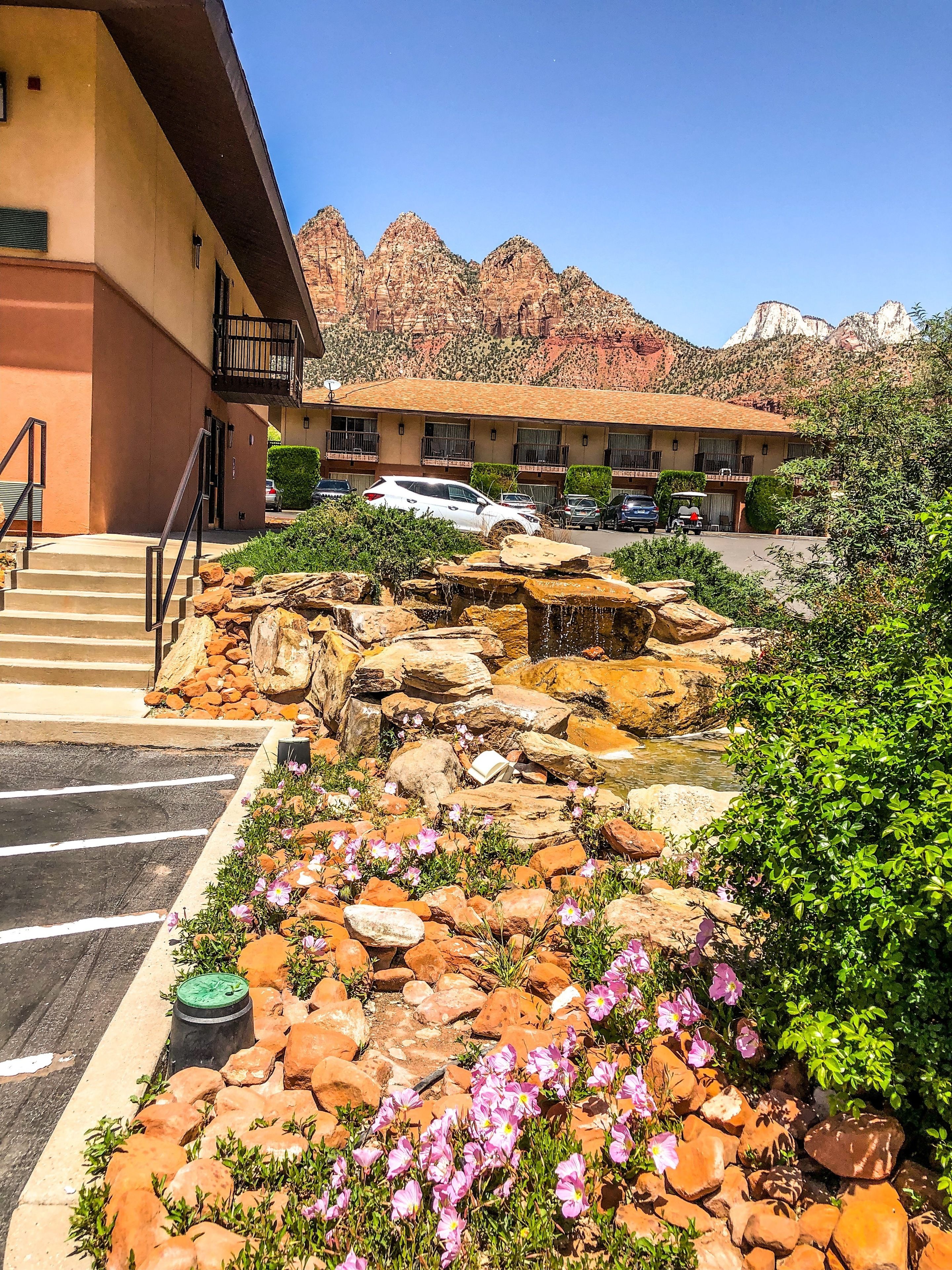 Un hotel con aparcamiento, un jardín de rocas con flores rosadas y montañas al fondo.