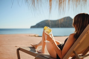 A person relaxing on a beach chair with a drink, overlooking a calm sea and a distant island.