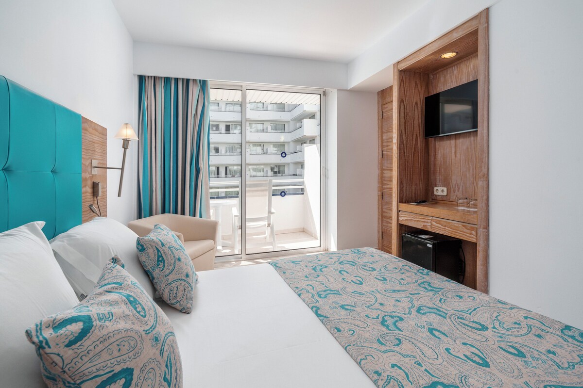 A hotel room with a bed, a sofa, a TV, and a balcony with a view of other buildings.