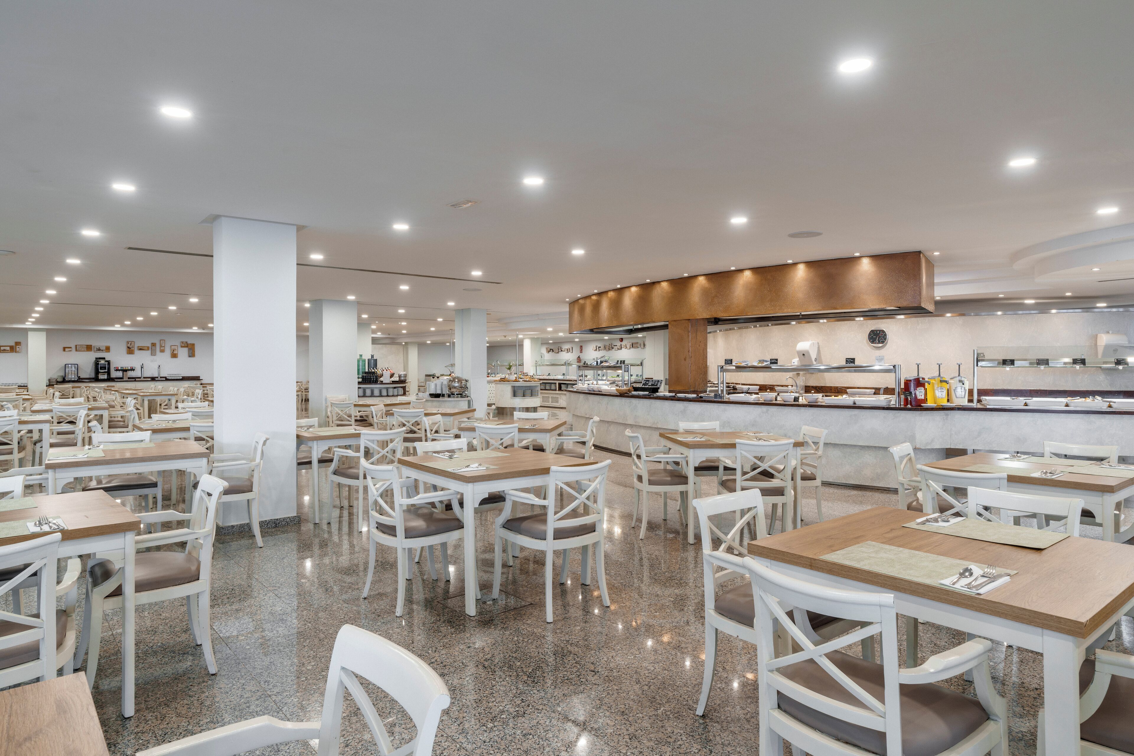 Une cafétéria spacieuse, dotée de nombreuses tables et chaises, d’un espace de service central et d’une cuisine moderne située à l’arrière.