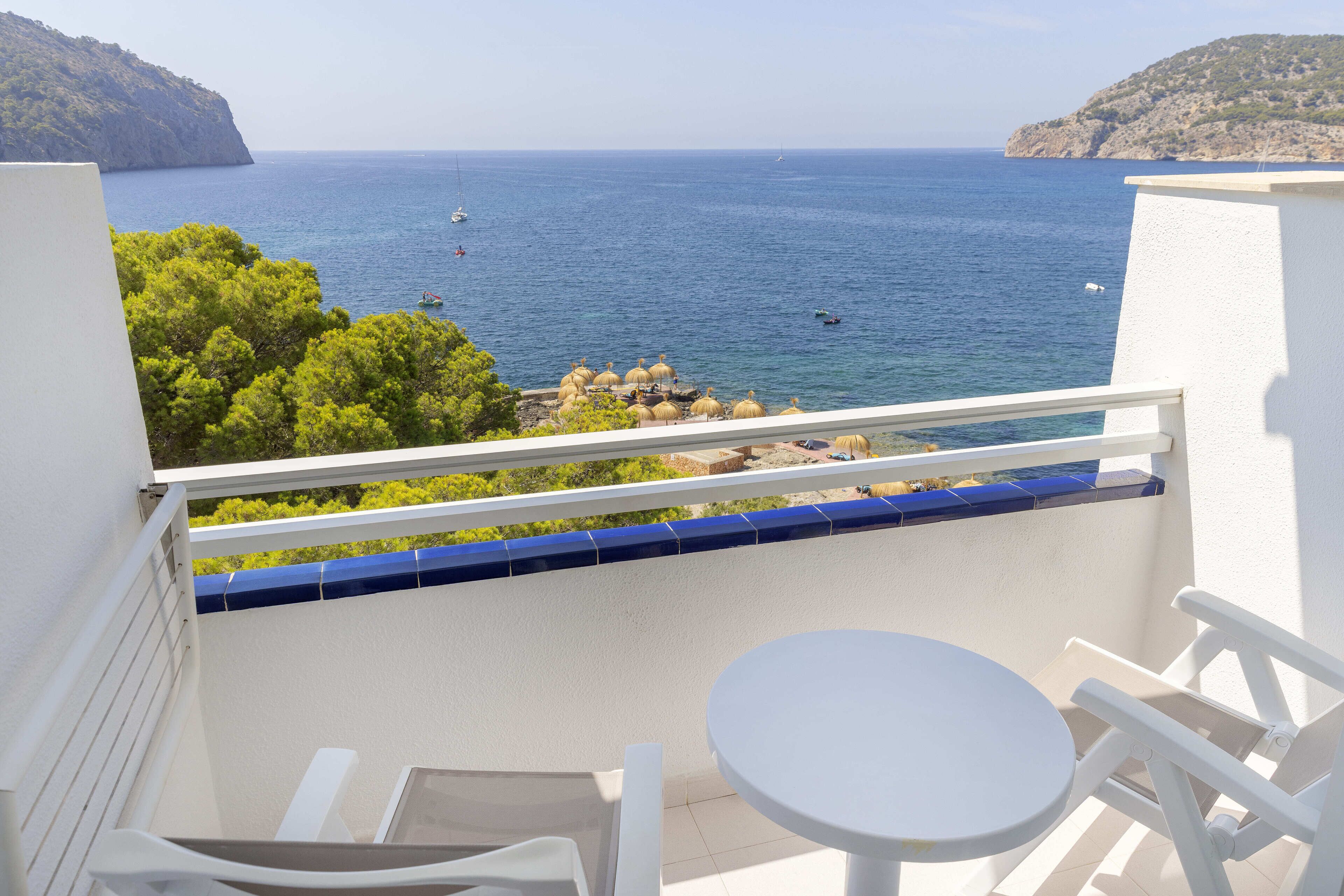Ein Balkon mit weißem Tisch und Stühlen, der Blick auf die Küste mit Booten und Grünflächen bietet.