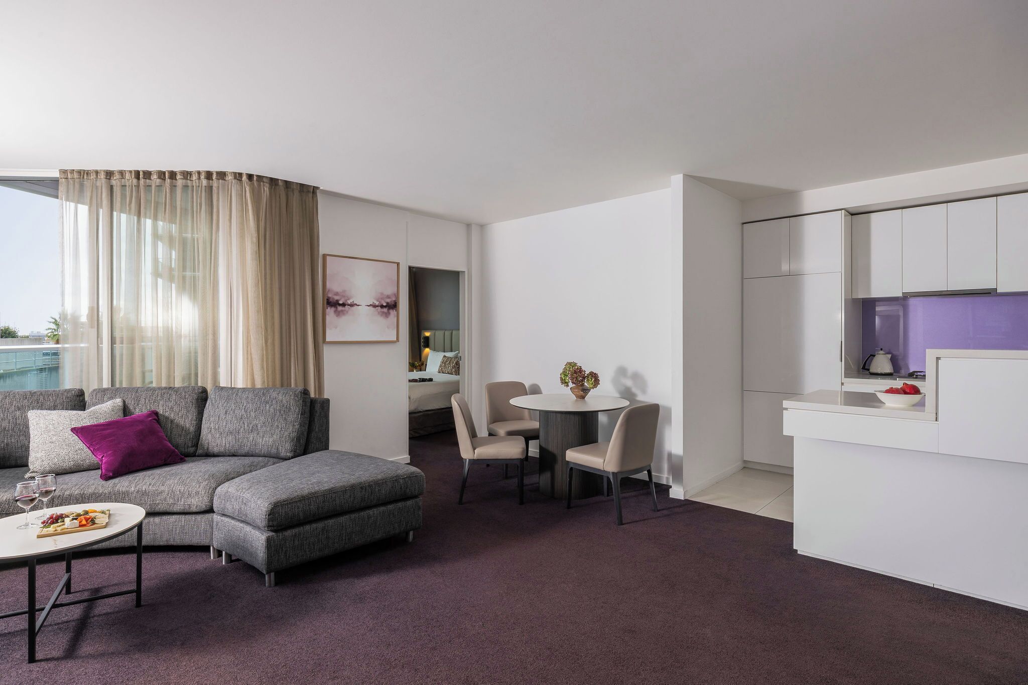 A modern hotel room with a grey sectional sofa, a dining table with chairs, a small kitchen area, and a view of the city.