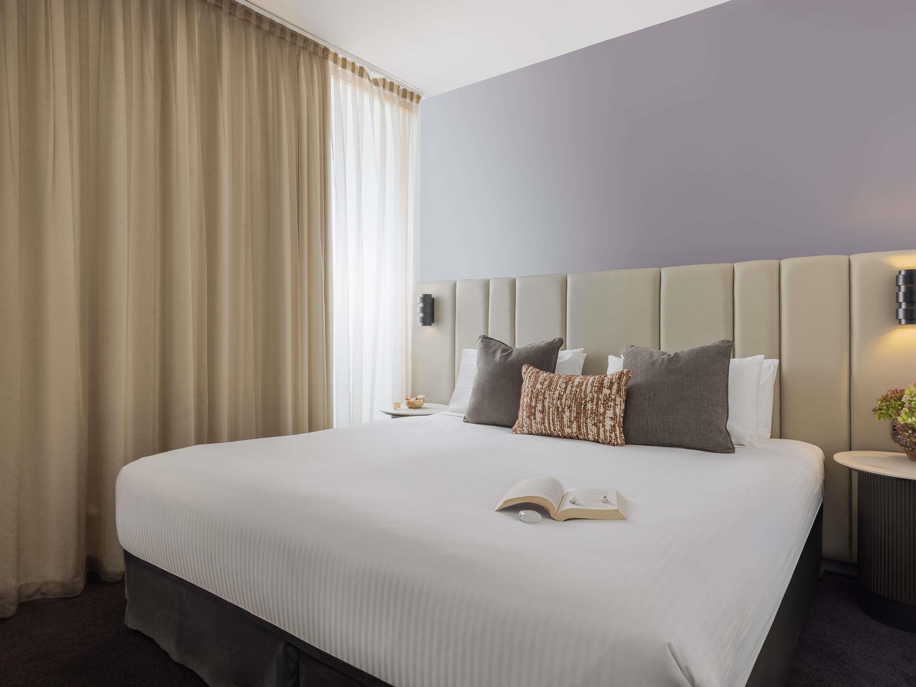 A hotel room with a large bed, a nightstand, a wall-mounted lamp, and a window with curtains.