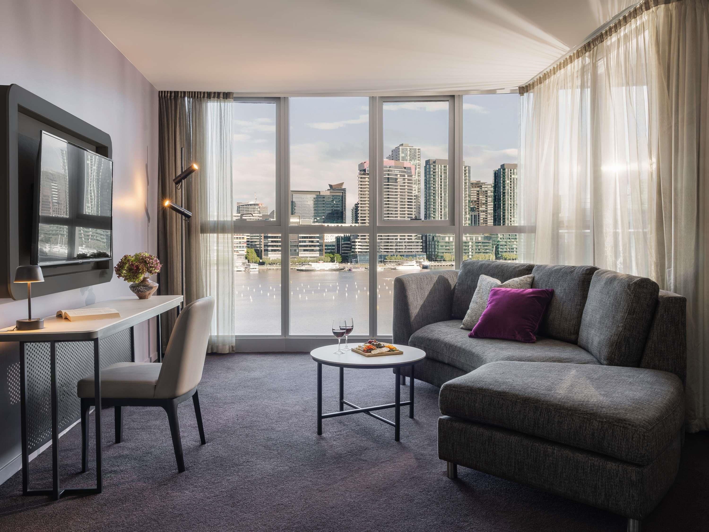 A modern hotel room with a large window, a flat-screen TV, a desk with a chair, a sofa, and a small coffee table.