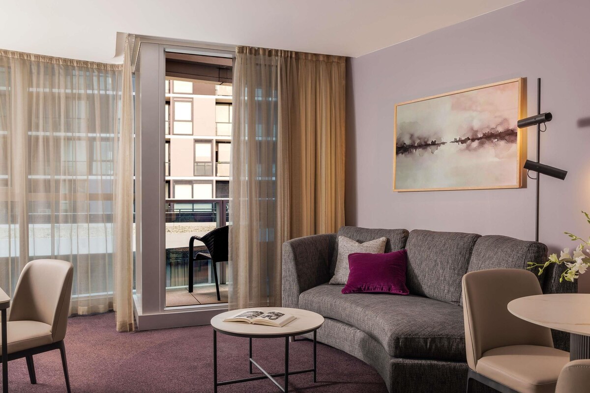 A modern living room with a grey sofa, a round coffee table, and a chair. There is a large window with sheer curtains, a painting on the wall, and a floor lamp.