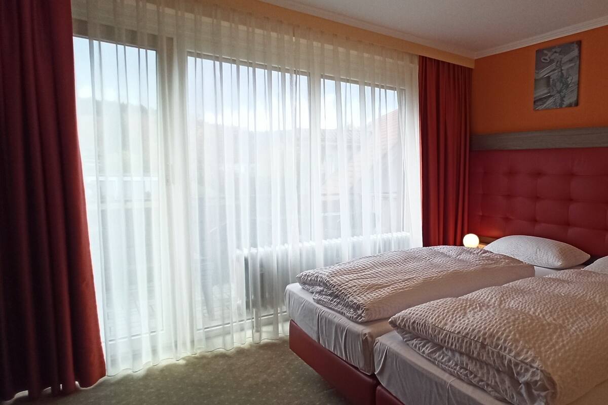 A hotel room with two beds, a red headboard, a window with sheer curtains, and a bedside table with a lamp.