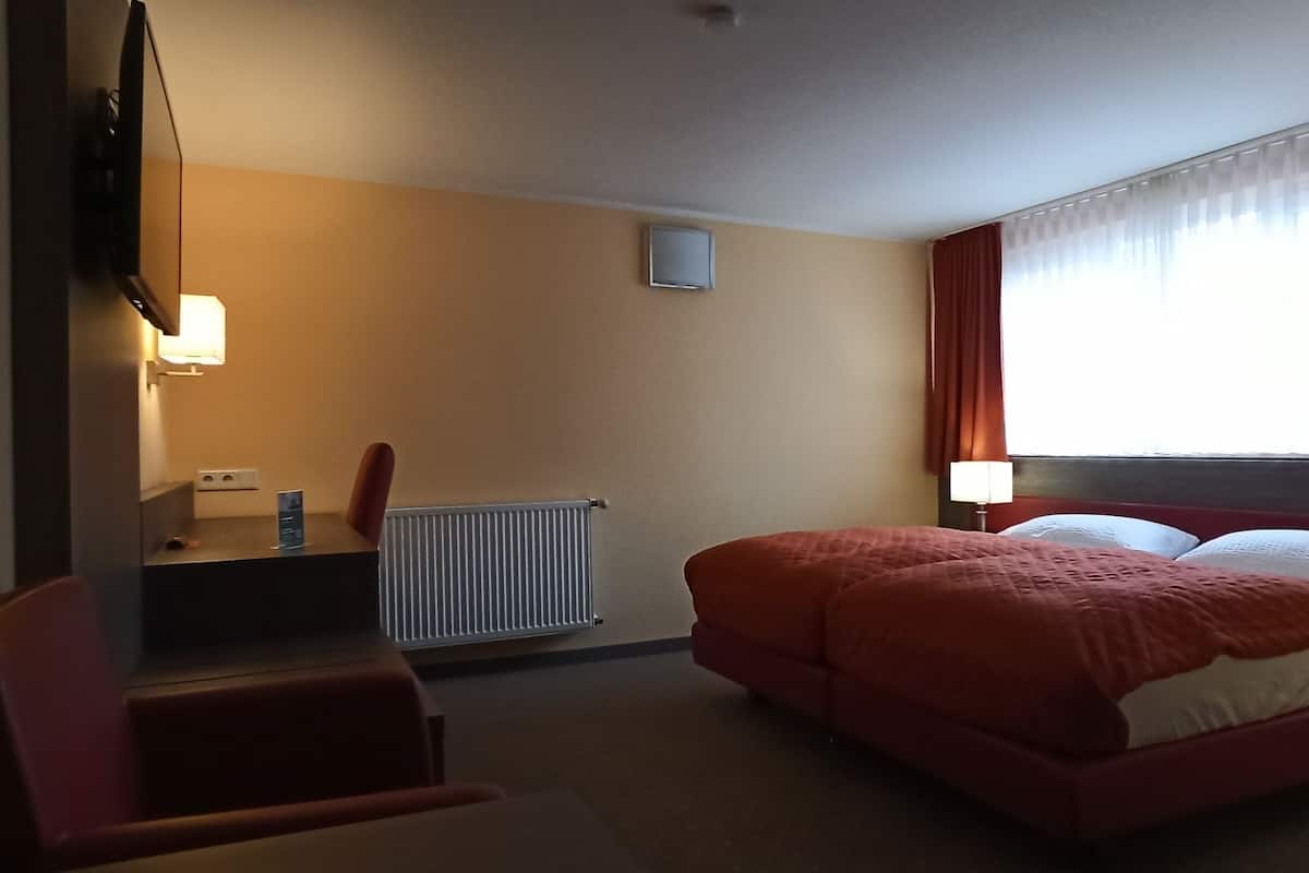 A hotel room with a bed, a desk, a chair, a radiator, and a window with curtains.