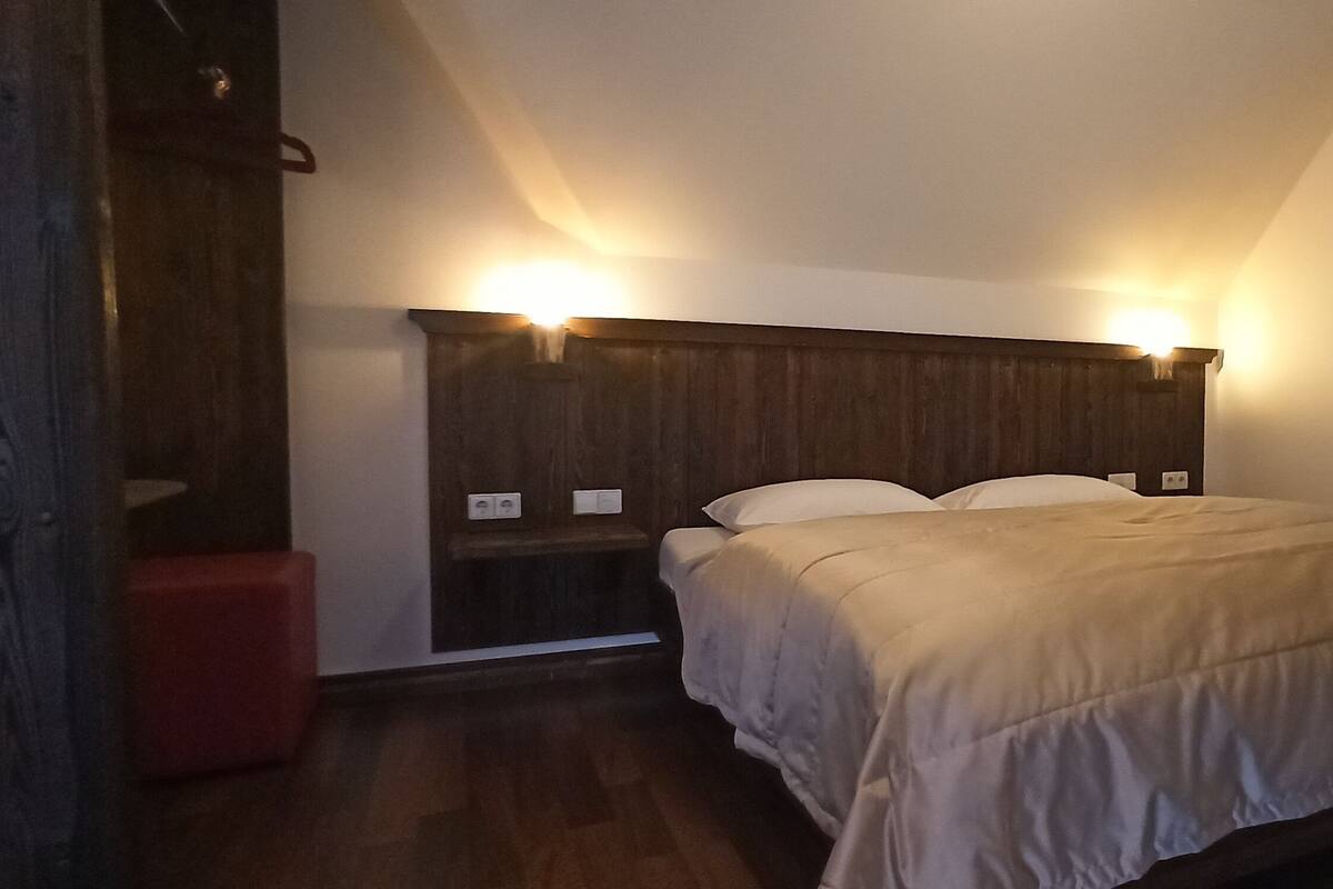 A bedroom with a wooden headboard, a bed with a beige blanket, a red bench, and a window with a view of a balcony.