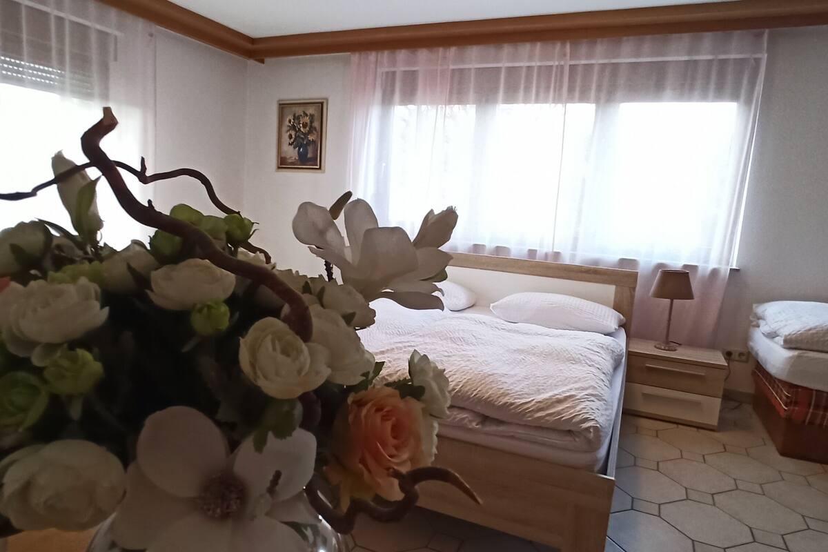 A bedroom with a bed, a bedside table, a nightstand, a window with curtains, and a floral arrangement on the table.