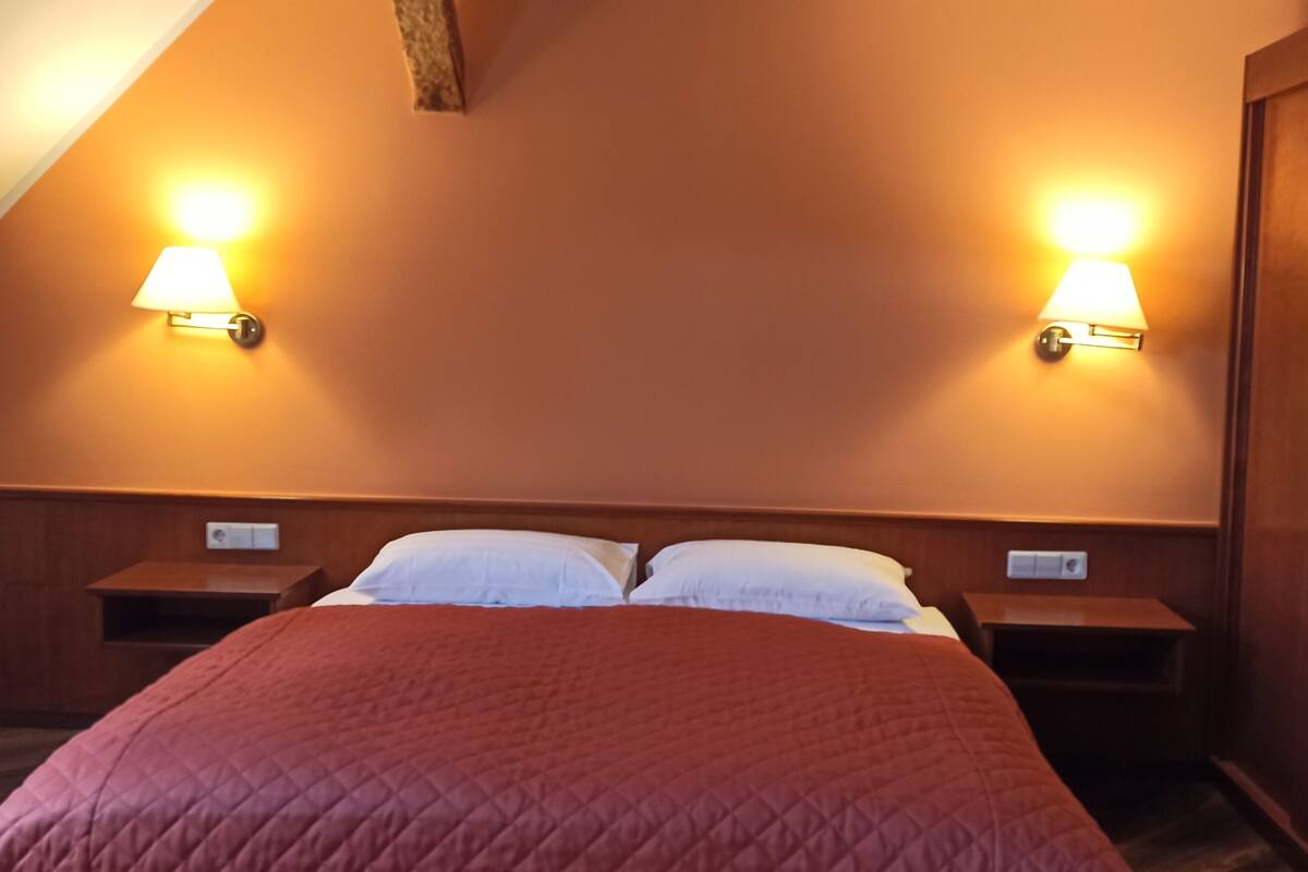 A bedroom with a sloped ceiling, two bedside lamps, a bed with a red quilt, and wooden headboard and nightstands.