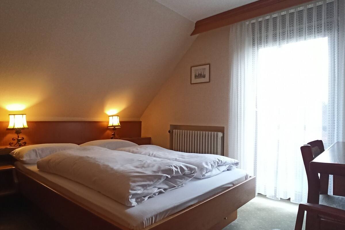 A hotel room with a bed, two bedside lamps, a radiator, a desk, and a chair.