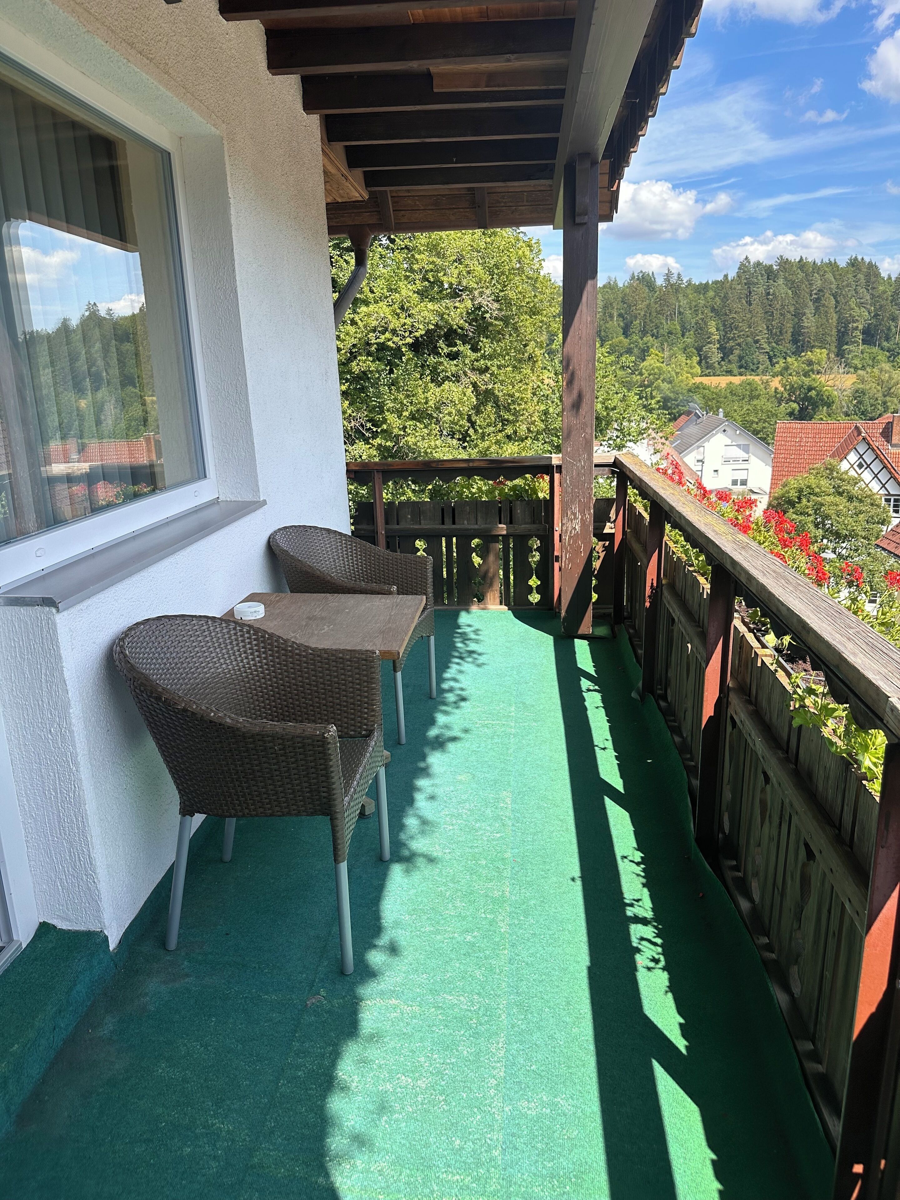 En balkong med ett golv av grön konstgräsmatta, flätade stolar och ett träräcke med utsikt över ett bostadsområde med träd och hus.