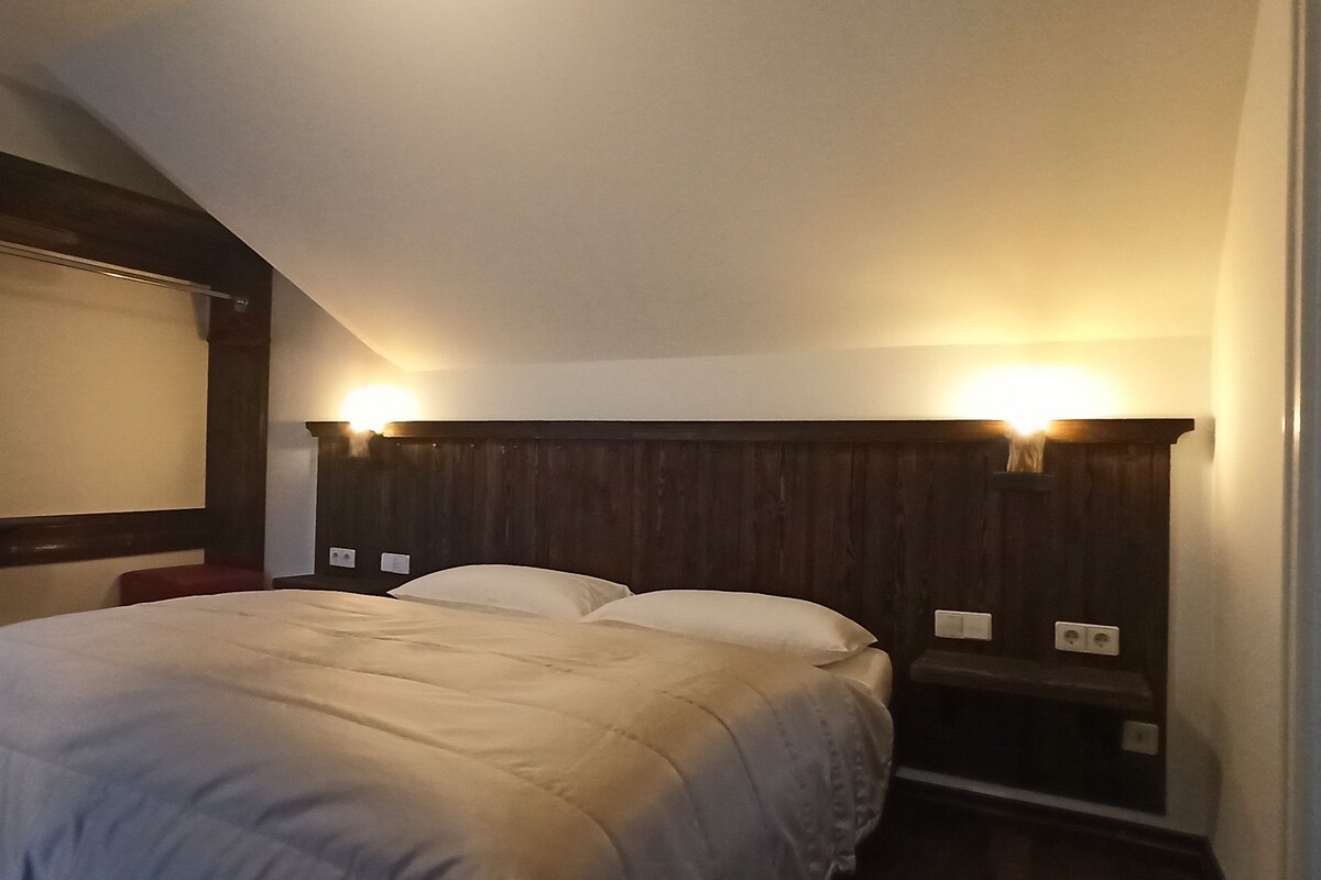 A bedroom with a large bed, wooden headboard, two bedside lamps, and a window with a blind.