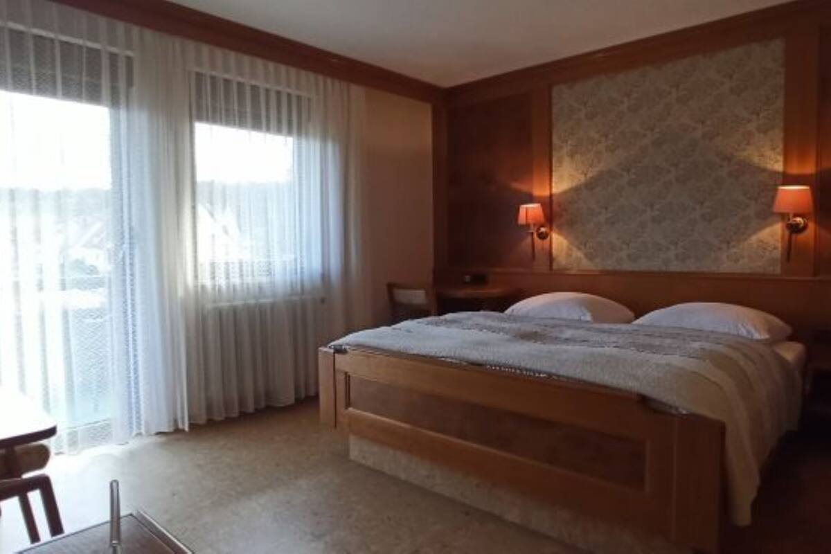 A hotel room with a wooden bed, two bedside lamps, a desk, and a window with sheer curtains.