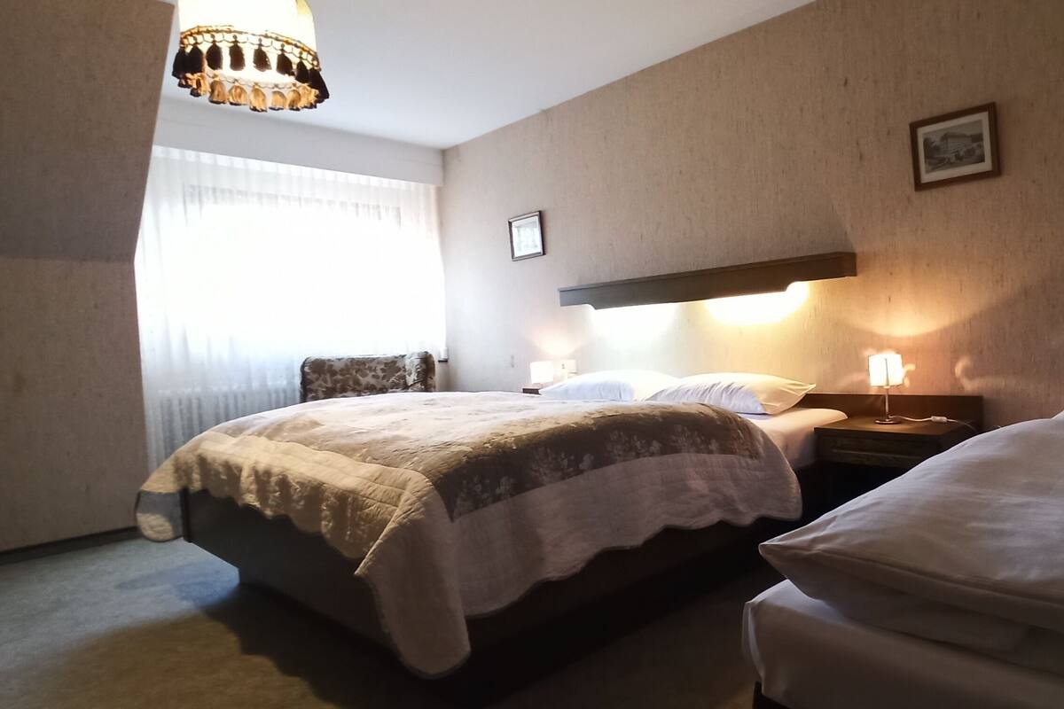 A hotel room with two beds, a large window with curtains, a chandelier, and a wall-mounted lamp.