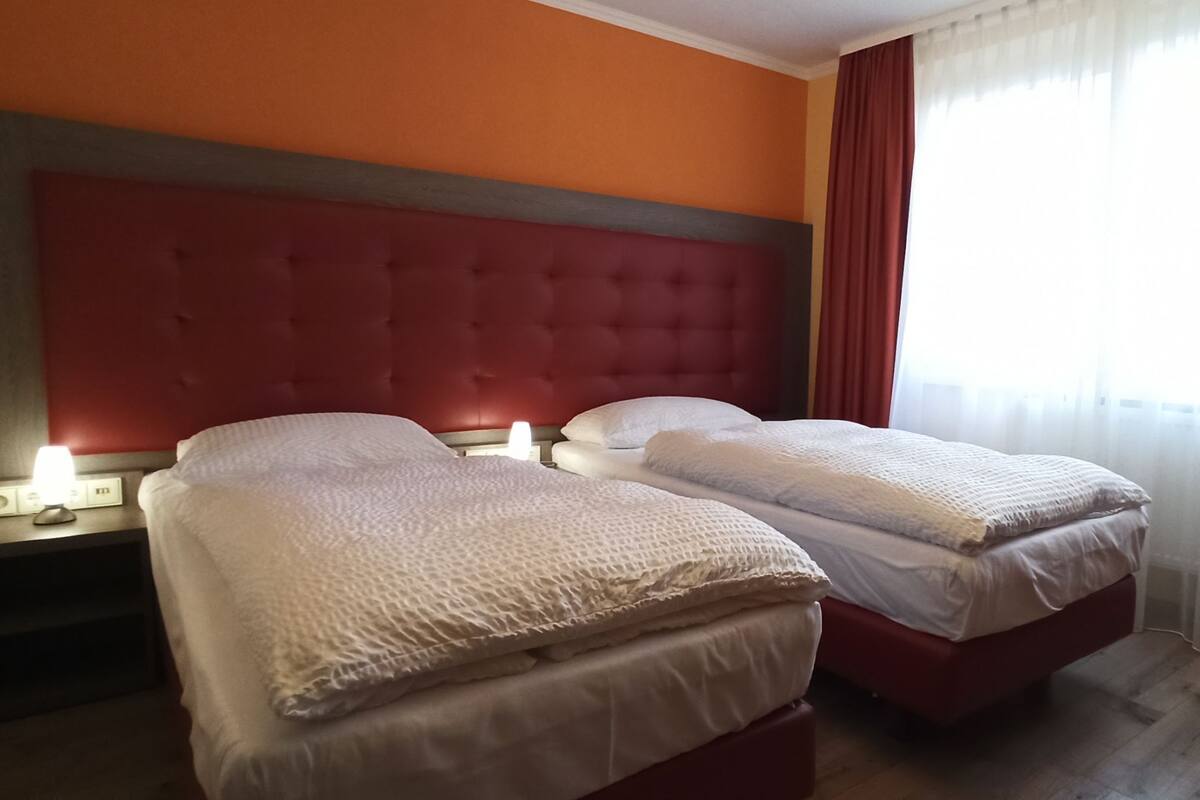 A hotel room with two beds, a red headboard, a nightstand with a lamp, and a window with curtains.