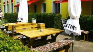 An outdoor seating area with wooden tables and benches, white umbrellas, and a yellow building with signage. - Hotel 4 Hufen (Schoenefeld)
