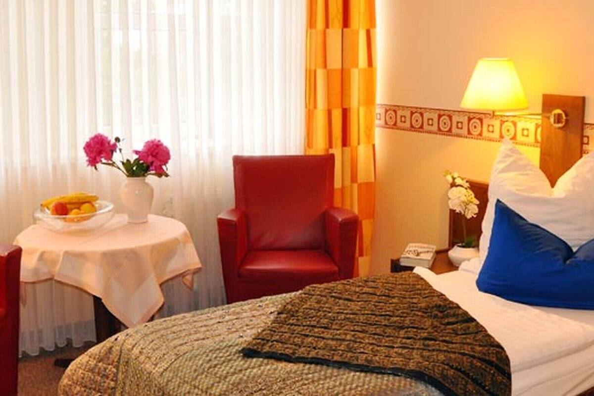 A hotel room with a bed, a red armchair, a small table with a fruit bowl, and a vase with flowers.