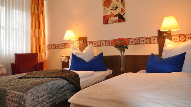 A hotel room with two beds, a red chair, a table with a vase of flowers, and a wall with a decorative pattern. - Hotel 4 Hufen (Schoenefeld)