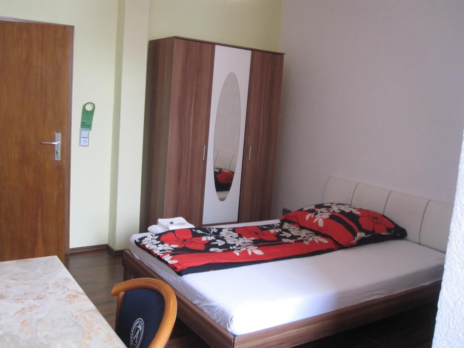 A bedroom with a bed, a wooden wardrobe, a small table, and a chair.