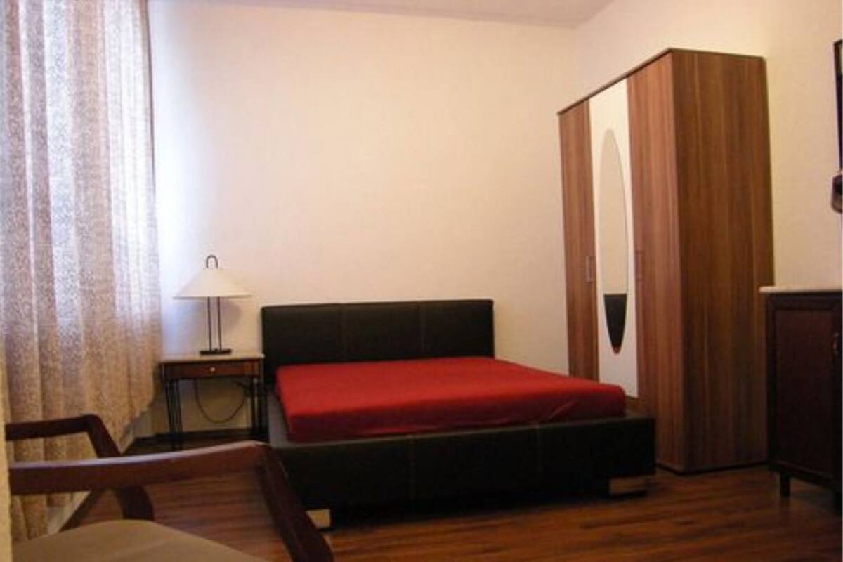 A bedroom with a bed, a wooden wardrobe, a desk with a lamp, and a chair.