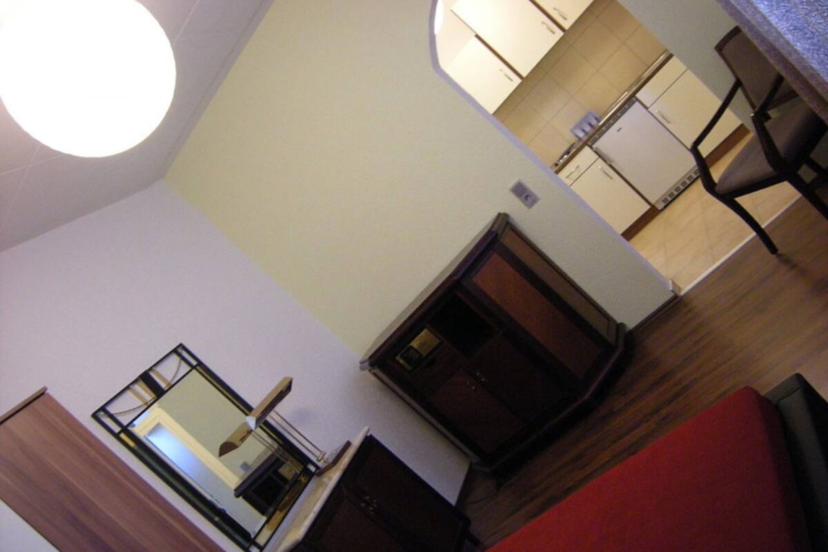 A room with a wooden floor, a red sofa, a small table, and a mirror. There is a kitchen area visible in the background.