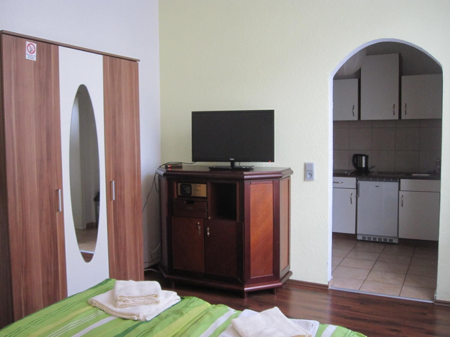 A room with a bed, a TV on a wooden cabinet, and a kitchen area visible through an archway.