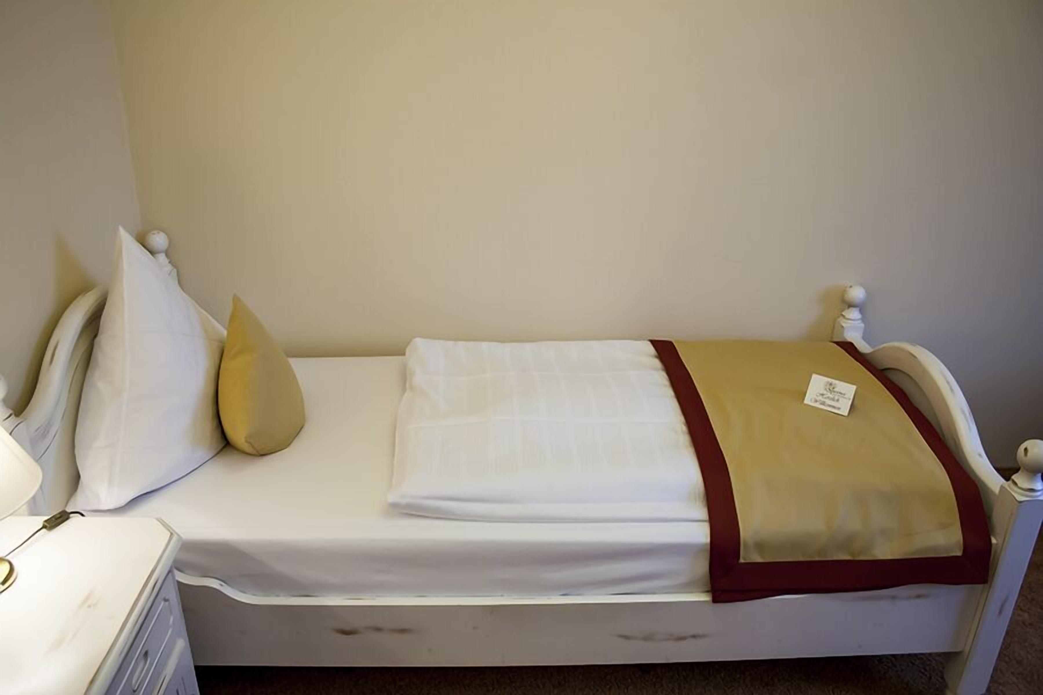 Una cama individual con colchón blanco, manta color beige y ribete rojo. Hay una etiqueta pequeña en la manta.