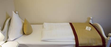 Una cama individual con colchón blanco, manta color beige y ribete rojo. Hay una etiqueta pequeña en la manta.