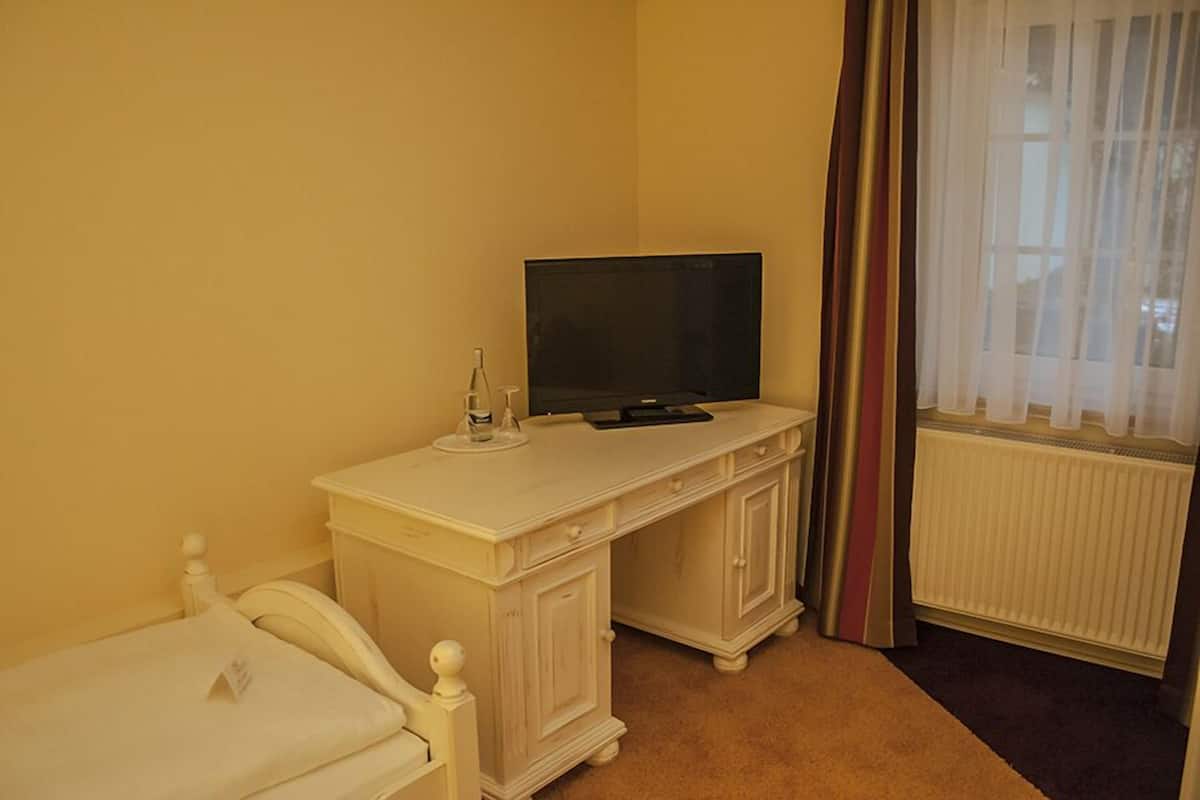A hotel room with a white bed, a desk with a television, a bottle, and a glass.