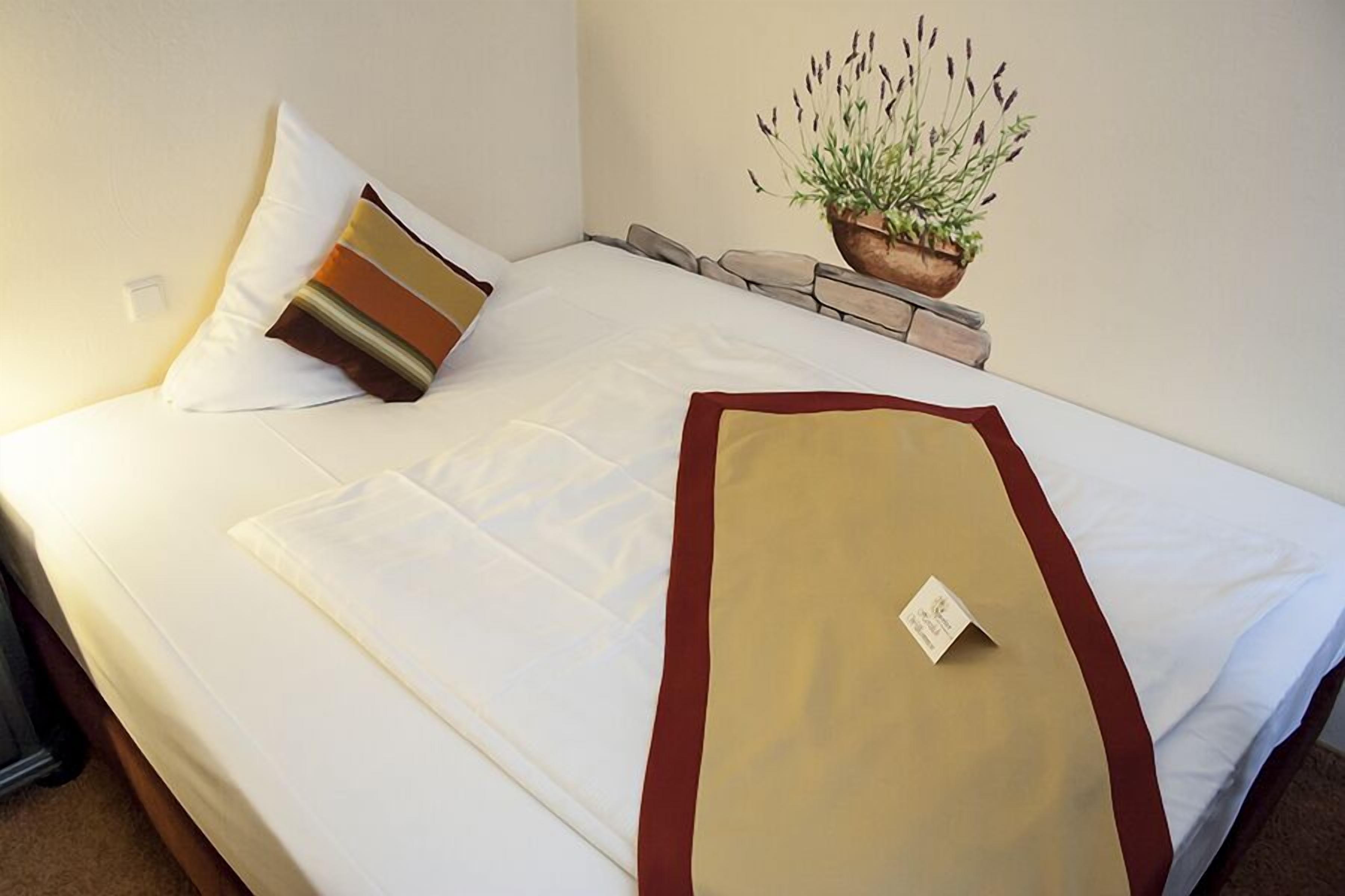 A neatly made bed with white linens, a brown and beige pillow, and a red and beige throw blanket. There is a small plant in a wooden pot on the nightstand beside the bed.