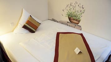 A neatly made bed with white linens, a brown and beige pillow, and a red and beige throw blanket. There is a small plant in a wooden pot on the nightstand beside the bed.