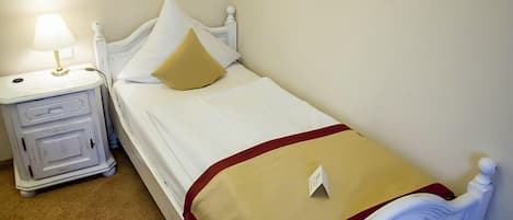 Uma cama de solteiro com cabeceira branca, uma mesinha de cabeceira com abajur e um cobertor bege.