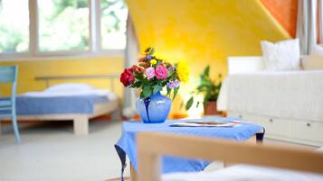 Een kamer met een bed, een blauwe tafel met een bloevas en een raam met uitzicht op groen.