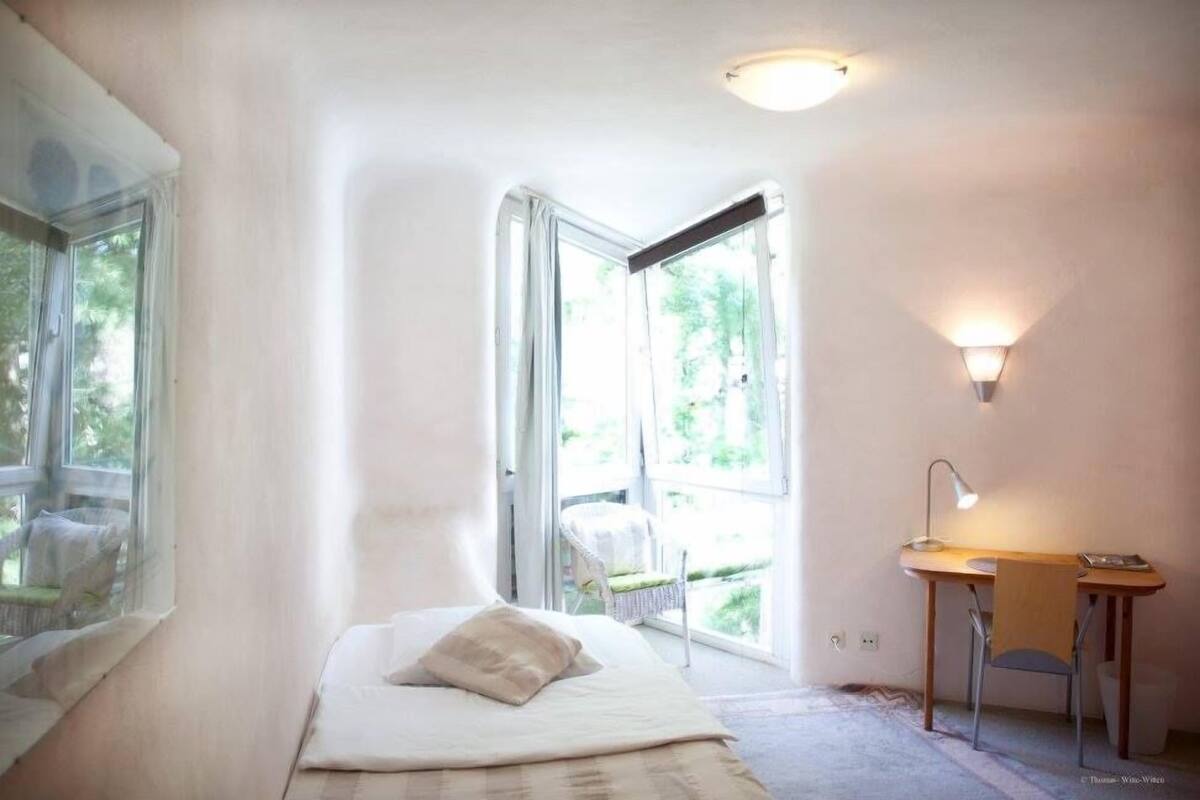 A bedroom with a bed, a desk, and a chair. There is a window with curtains and a view of greenery outside.
