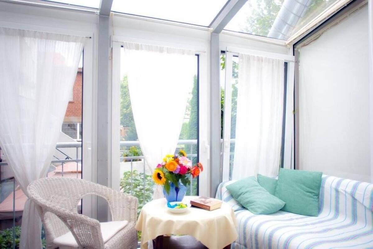 A sunlit room with a wicker chair, a striped sofa, and a table with a vase of flowers.