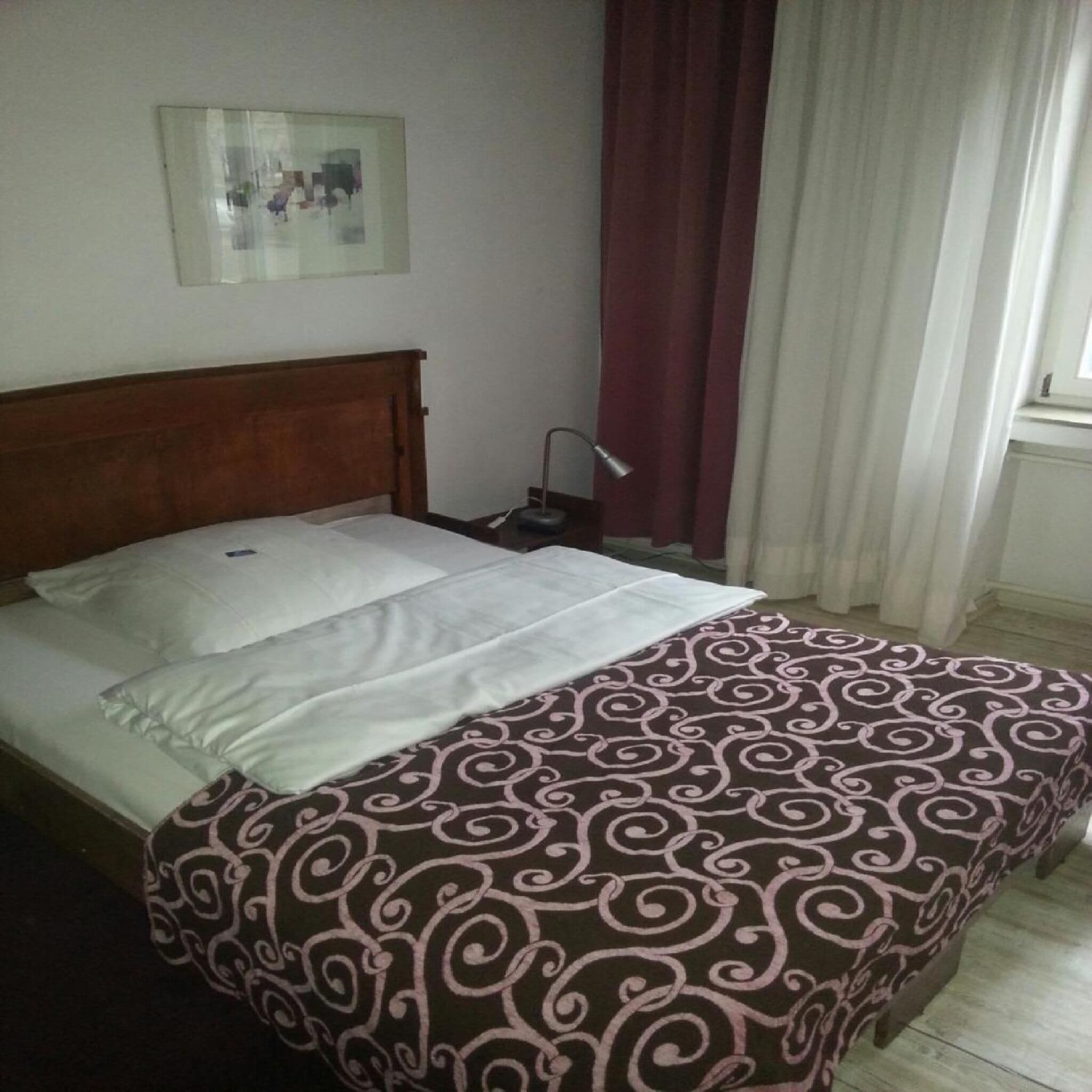 A bed with a patterned bedspread, a wooden headboard, a bedside table with a lamp, and a window with curtains.