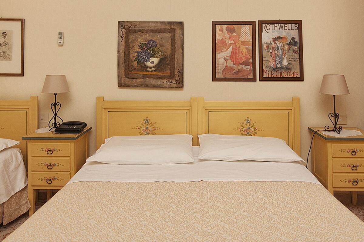 Habitación de hotel con dos camas individuales y una cama más grande, cada una con mesitas de noche y lámparas a juego. En la pared, hay cuadros enmarcados y un cartel publicitario de chocolate con leche.